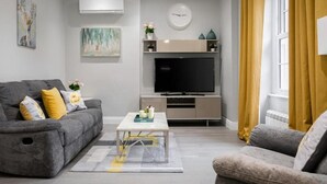 A modern living room with a grey sofa, a coffee table, a flat-screen TV, and a yellow curtain. - Superior 3 Bedroom Apartment (London)