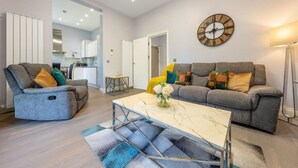 A living room with a sofa, armchair, coffee table, and a clock on the wall. - Deluxe 2 Bedroom Apartment (London)