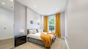 A modern bedroom with a bed, bedside table, and a window with curtains. - Deluxe 2 Bedroom Apartment (London)