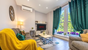 A modern living room with a sofa, a coffee table, a TV, and a window with curtains. - Deluxe 2 Bedroom Apartment (London)