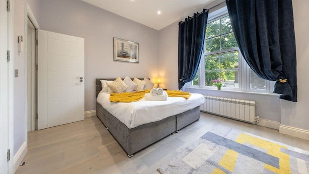 A bedroom with a bed, a radiator, a window with curtains, and a door. - Deluxe 2 Bedroom Apartment (London)