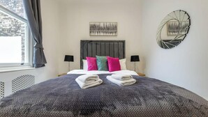 A neatly made bed with pillows and folded towels, a wall-mounted abstract art piece, and a window with curtains. - Victorian Style One-bedroom Apartment (London)