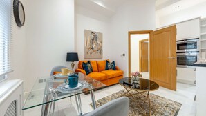 A modern living room with a glass dining table, a sofa with orange cushions, a painting on the wall, and a kitchen in the background. - Victorian Style One-bedroom Apartment (London)