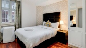 A hotel room with a bed, a nightstand, a lamp, a mirror, a window with curtains, and a radiator. - Superior Three Bedroom Apartment Opposite Harrods (London)