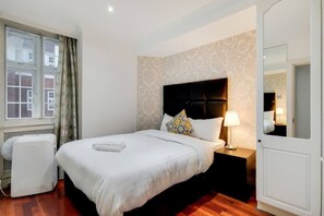 Ein Hotelzimmer mit Bett, Nachttisch, Lampe, Spiegel, Fenster mit Vorhängen und Heizkörper.