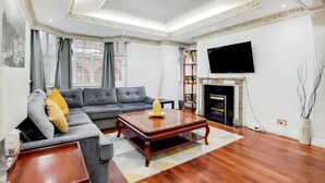 A living room with a sofa, a coffee table, a fireplace, and a television mounted on the wall. - Superior Three Bedroom Apartment Opposite Harrods (London)