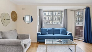 Un salon avec un canapé bleu, un fauteuil gris, une table basse en verre et une vue sur les immeubles par la fenêtre.