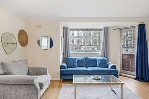 Ein Wohnzimmer mit einem blauen Sofa, einem grauen Sessel, einem Glastisch und Blick auf Gebäude durch ein Fenster.