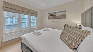 Ein Schlafzimmer mit einem großen Bett, einem Fenster mit Blick auf Gebäude, einem Gemälde an der Wand und einem Nachttisch mit einer Lampe.