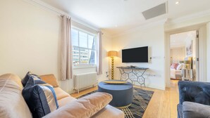 A modern living room with a sofa, ottoman, and a television on a stand. - Deluxe One Bedroom Apartment (London)