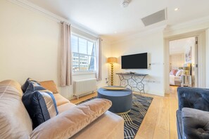 A modern living room with a sofa, ottoman, and a television on a stand. - Deluxe One Bedroom Apartment (London)
