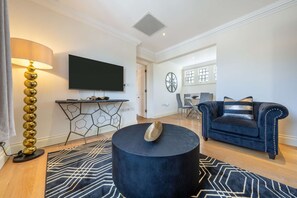 A living room with a round black coffee table, a blue sofa, a TV mounted on the wall, and a dining area in the background. - Deluxe One Bedroom Apartment (London)