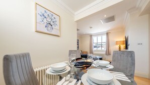A dining area with a glass table set for four, surrounded by grey upholstered chairs. A framed floral artwork is on the wall, and a television is mounted on the wall to the right. - Deluxe One Bedroom Apartment (London)