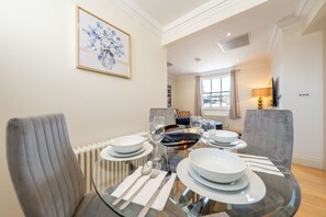 A dining area with a glass table set for four, surrounded by grey upholstered chairs. A framed floral artwork is on the wall, and a television is mounted on the wall to the right. - Deluxe One Bedroom Apartment (London)