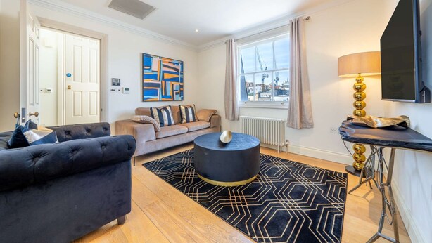 A modern living room with a sofa, a round black coffee table, a TV mounted on the wall, and a view of the city through a large window. - Deluxe One Bedroom Apartment (London)