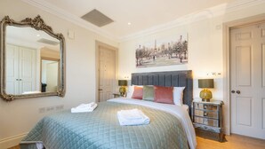 A hotel room with a large mirror, a bed with a quilted bedspread, two bedside lamps, a nightstand, and a painting of a park scene. - Deluxe One Bedroom Apartment (London)