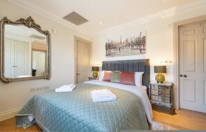 A hotel room with a large mirror, a bed with a quilted bedspread, two bedside lamps, a nightstand, and a painting of a park scene. - Deluxe One Bedroom Apartment (London)