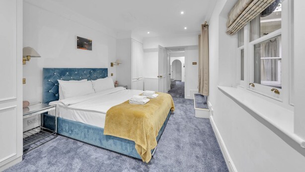 Una camera d'albergo con un letto, un comodino, una finestra con tende e un corridoio che conduce a un'altra stanza.