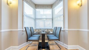 A dining area with a table set for four, surrounded by chairs, in a room with large windows and wall-mounted lighting. - Exclusive 2-bedroom Apartment (London)