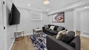 A modern living room with a grey sofa, a flat-screen TV mounted on the wall, a small side table, and a patterned rug. - The Windsor Mainstay (Windsor)