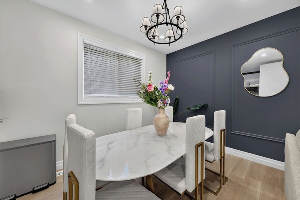 A dining area with a marble table, white upholstered chairs, a vase with flowers, a chandelier, a window with blinds, and a mirror on the wall.