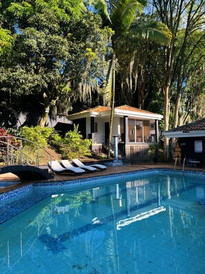 Piscina com espreguiçadeiras, um alpendre coberto e uma casa rodeada de árvores.