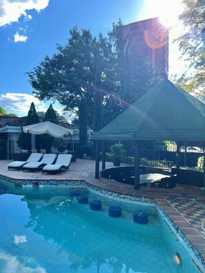 Ein Poolbereich mit Liegestühlen, einer überdachten Lounge und einer Poolkante mit Fliesen.