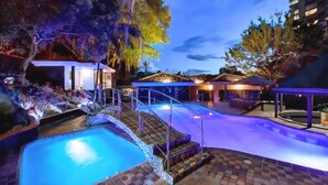 Zona de piscina con fuente de agua, escalones que llevan a un área de estar techada y un edificio con tejado de baldosas.