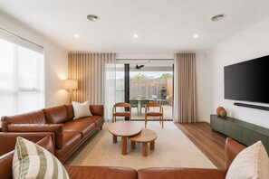Ein Wohnzimmer mit einer Couch, zwei Sesseln, einem Couchtisch, einem Fernseher und einer Glastür zum Garten.