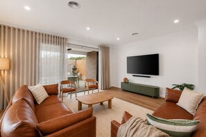 Ein modernes Wohnzimmer mit einer Ledersofa, einem Couchtisch, einem an der Wand montierten Fernseher und einem Essbereich, der durch eine Glastür sichtbar ist.