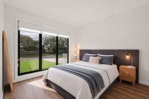 Ein Schlafzimmer mit einem großen Schiebeglasfenster zum Rasen, einem Bett mit gestreifter Bettwäsche und einem Nachttisch mit einer Lampe.