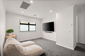 Ein modernes Wohnzimmer mit Fernseher, einer Zimmerpflanze, einem beigefarbenen Sofa und einem Fenster mit Jalousien.