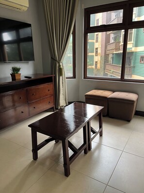A room with a wooden table, a sofa, a TV, a window with a view of buildings, and a curtain. - One Bedroom Mountain Respite at Crosswinds (Tagaytay City)
