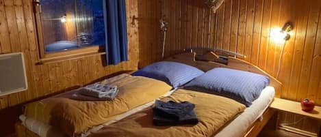 A wooden cabin room with a bed, a window with curtains, and a wall-mounted light fixture.