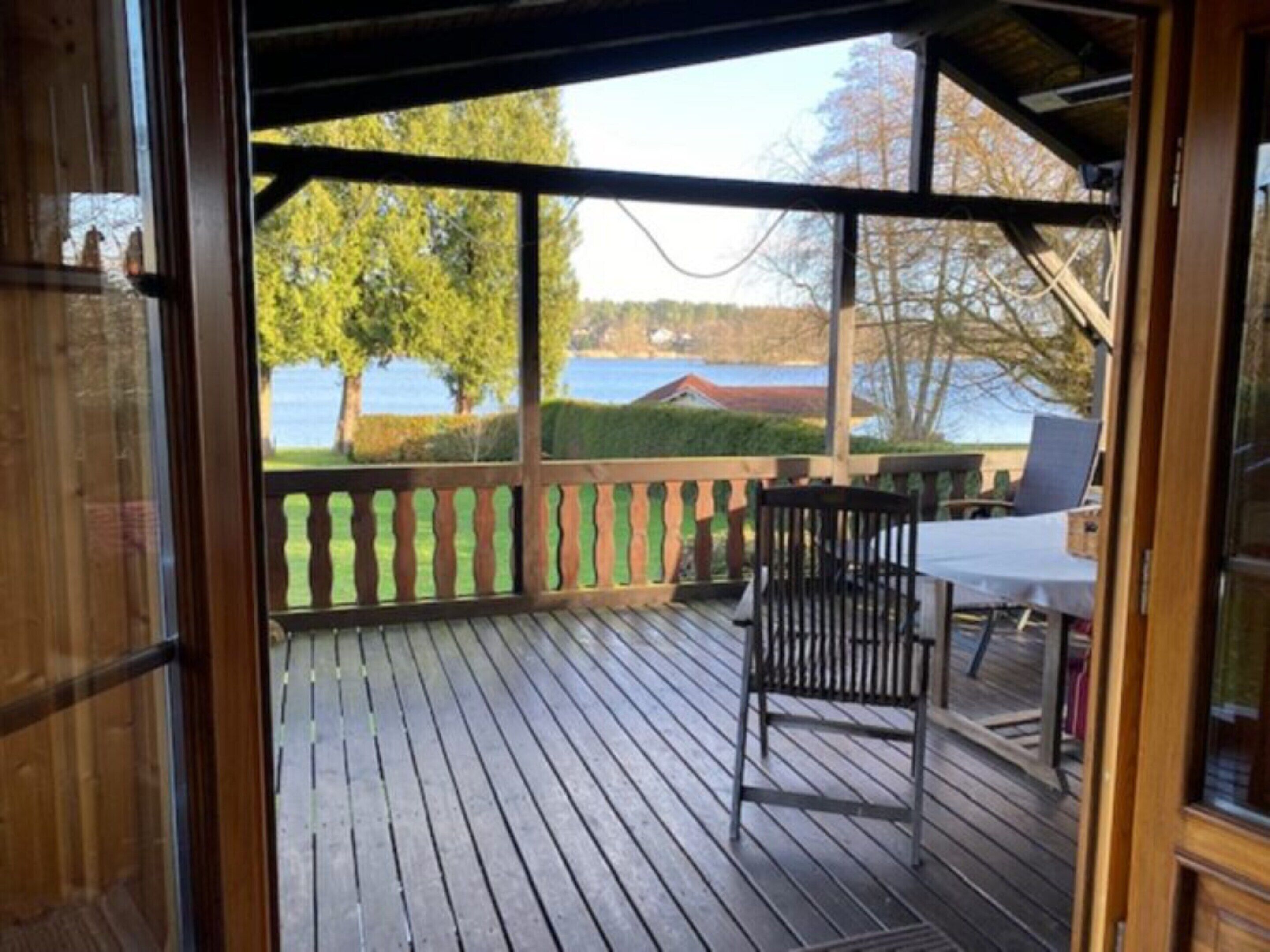 A wooden deck with a table and chairs, overlooking a lake with trees and a house in the background.