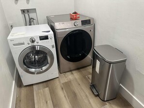 A laundry area with a front-loading washer and dryer, a stainless steel trash bin, and a wooden floor.