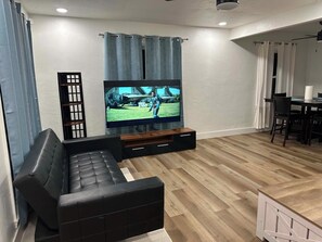 Een moderne woonkamer met een zwarte leren bank, een flatscreen televisie bevestigd aan een houten tv-meubel, en een eethoek met een tafel en stoelen.