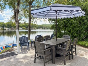 An outdoor patio with a table and chairs, a striped umbrella, and a view of a lake. - Lakefront Escape - Dock, Beach, Grill & Hot Tub (Milton)