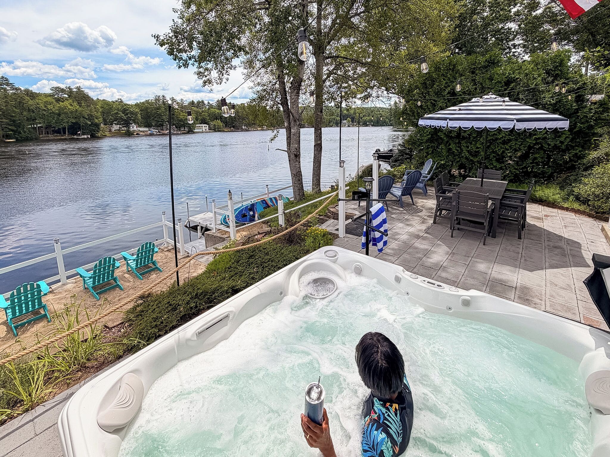 A hot tub with a person using a smartphone, surrounded by outdoor seating and a view of a lake.