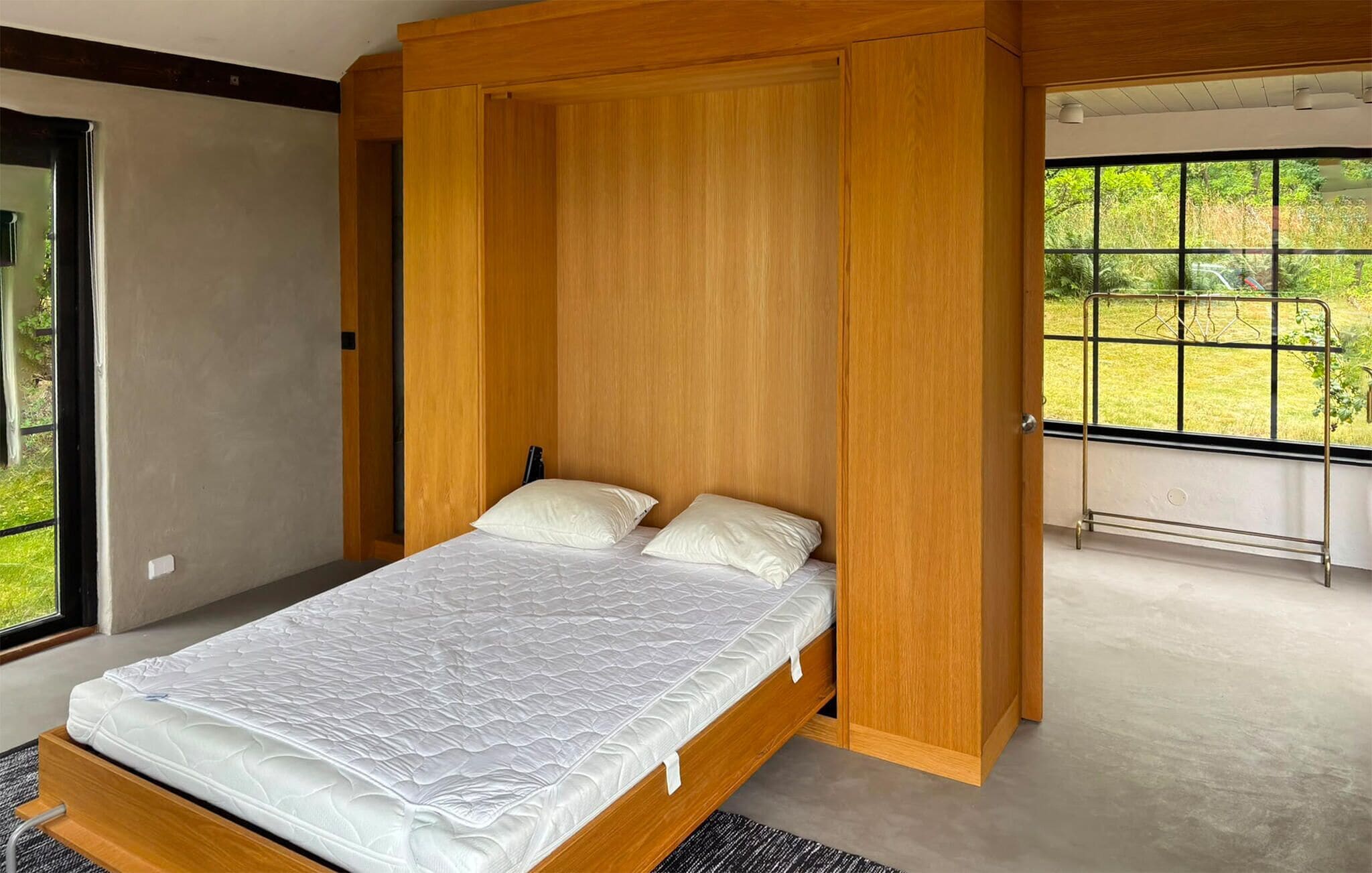 Een moderne slaapkamer met een houten bed, een groot raam met uitzicht op groen en een balkon met een metalen hekwerk.