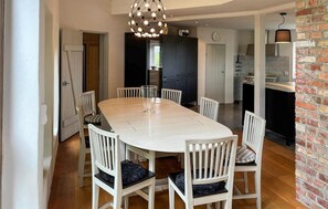 A dining area with a white table and chairs, a hanging light fixture, and a kitchen in the background. - 5 bedroom luxurious home in Simrishamn (Simrishamn)