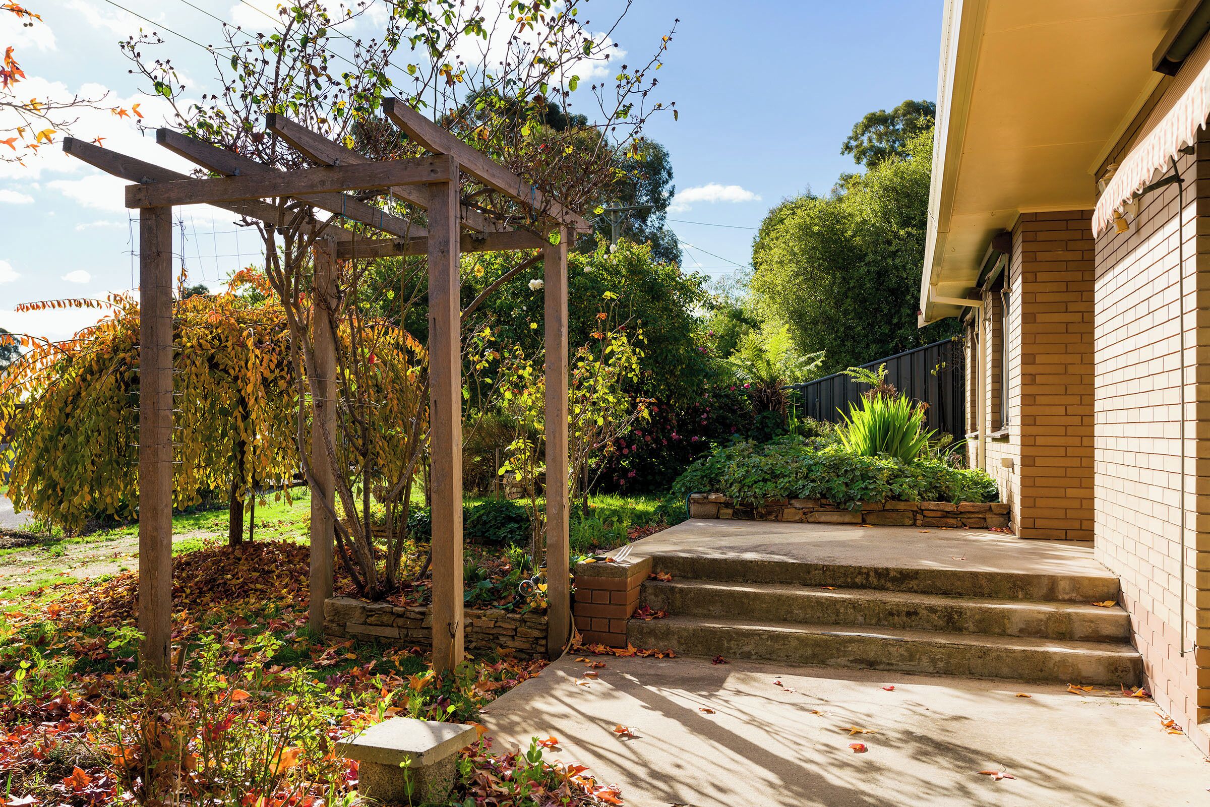 Un jardin agrémenté d’une pergola en bois, d’un escalier en pierre menant à un bâtiment en briques, ainsi que de diverses plantes et arbres.