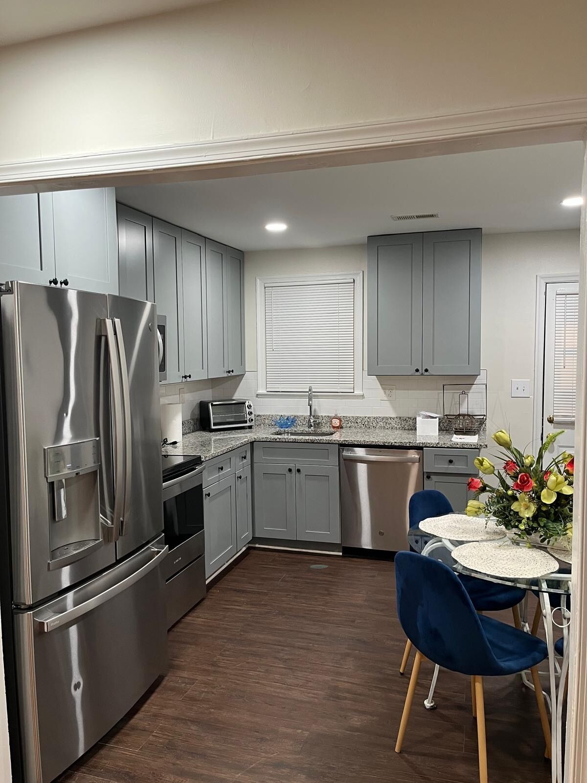 A modern kitchen with grey cabinets, stainless steel appliances, a dining area with blue chairs, and a window with blinds.