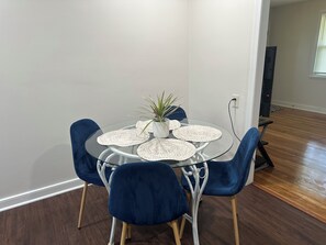 A round glass table with white placemats, a potted plant, and blue chairs in a room with hardwood flooring and white walls. - Charming little house (Richmond)