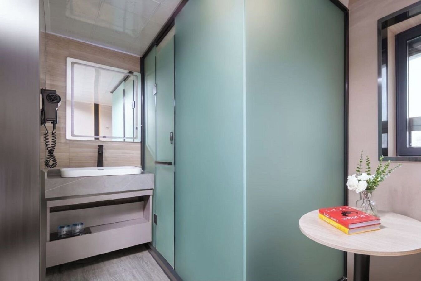 Baño compacto con lavamanos, espejo y una mesita con un libro y flores.