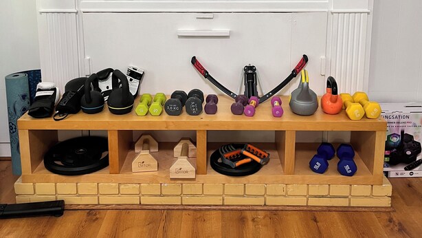 A wooden shelf with various fitness equipment, including dumbbells and kettlebells, against a white wall.