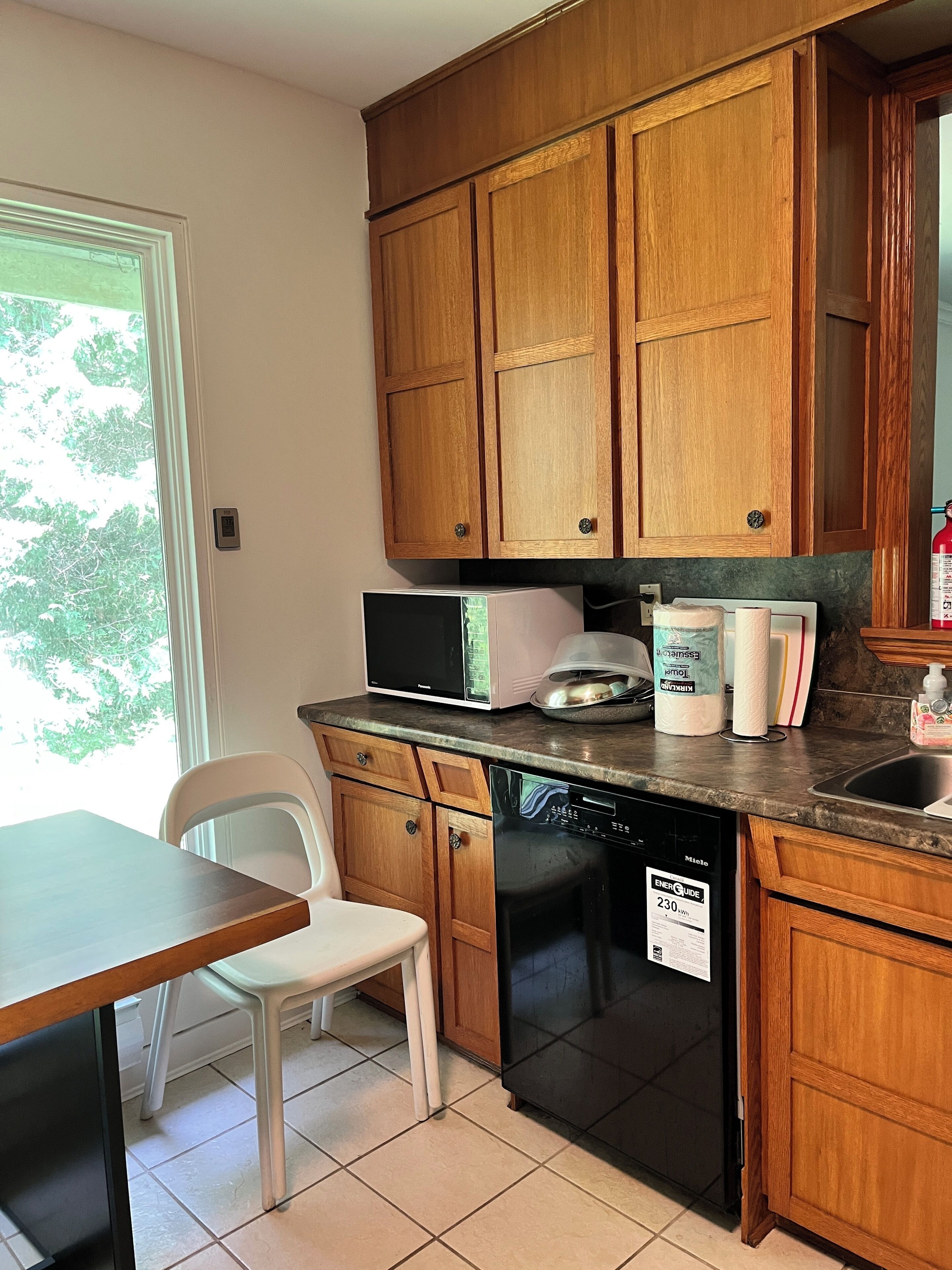A kitchen with wooden cabinets, a microwave, a toaster, a dishwasher, and a window with a view of trees.