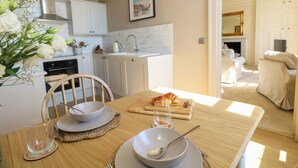 A modern kitchen with white cabinets, a dining table set with plates, bowls, and cutlery, and a view into a living area with a sofa and a fireplace. - 123 On The Sea Victorias Place (Weymouth)