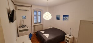 Ein Schlafzimmer mit Bett, Schreibtisch, Stuhl, Kommode, Heizkörper, einem Fenster mit Fensterläden, einer Pendelleuchte und Wandbildern.