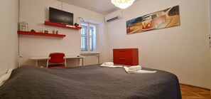 Ein Schlafzimmer mit Bett, Schreibtisch, Stuhl, Fernseher, Heizkörper, Fenster und einem Gemälde.